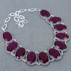 Könnte beinhalten: Eine Statement-Halskette mit zehn großen, ovalen, tiefvioletten facettierten Steinen. Jeder Stein ist in einer dekorativen silberfarbenen Metallfassung mit gedrehter Kordelbordüre gefasst. Die Kette hat eine silberfarbene Gliederkette.