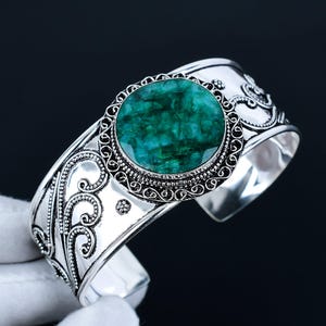 Può includere: Braccialetto rigido in argento con un designo ornato e una grande pietra preziosa rotonda verde al centro.