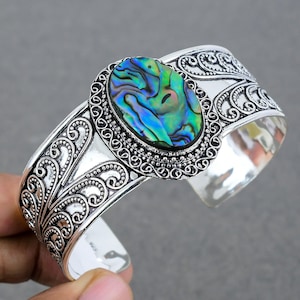 May include: A silver cuff bracelet with an oval abalone shell centerpiece. The shell displays iridescent blues, greens, and purples. The bracelet has an open design with ornate scrollwork and a textured finish, creating a vintage aesthetic.