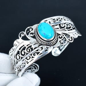 May include: A silver cuff bracelet with an oval turquoise stone centerpiece. The bracelet features intricate openwork designs and a textured border around the stone. The turquoise stone is a vibrant blue color.
