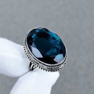 May include: A ring featuring a large, oval-cut, deep blue gemstone set in a decorative silver band. The metal setting displays intricate beaded and etched detailing around the perimeter of the stone, providing a textured finish to the silver frame.