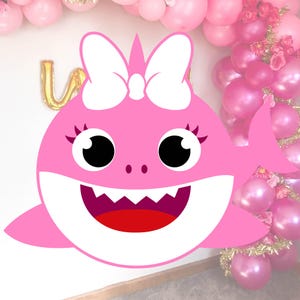 May include: Pink baby shark with a white bow, big eyes, and a wide open mouth. The shark is against a white background with pink and gold balloons.
