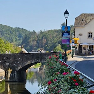 Könnte beinhalten: Eine Steinbrücke über einen Fluss in einer kleinen Stadt. Die Brücke hat zwei Bögen und ist mit Blumen bepflanzt. Auf der rechten Seite des Bildes befindet sich ein Café namens "Cafe du Pont".