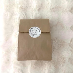 K&ouml;nnte beinhalten: Ein brauner Papiersack, verschlossen mit einer d&uuml;nnen Schnur, mit einem runden Aufkleber mit dem Text "ellashamstertreats" und einer Hamsterillustration. Der Beutel liegt auf einem flauschigen, wei&szlig;en Hintergrund.