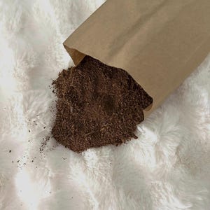 May include: A brown paper bag partially open, spilling out a pile of dark brown, finely ground material. The bag is set against a fluffy, white, textured background, creating a contrast between the rough and smooth textures.