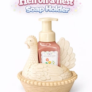 以下が含まれることがあります： クリーム色のニワトリ型のソープホルダーとピンク色のソープディスペンサー。ソープディスペンサーには「Bath & Body Works」のラベルが付いています。ホルダーは巣の形をしており、上部に「Hen on a nest Soap Holder」の文字があります。