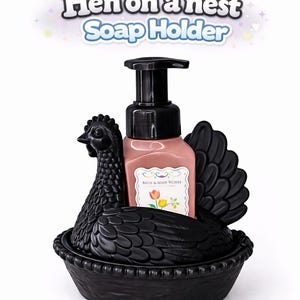以下が含まれることがあります： ピンク色の石鹸ディスペンサーが付いた、黒い鶏の形をした石鹸ホルダー。石鹸ディスペンサーには黒いポンプと花の模様のラベルが付いています。ホルダーは巣の中の鶏の形をしています。画像には「Hen on a nest Soap Holder」と書かれています。