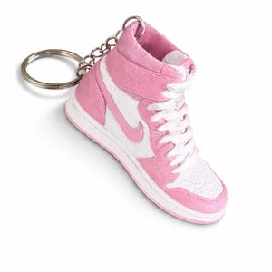 May include: A miniature high-top sneaker keychain featuring a pink and white color scheme. The shoe has a pink suede-like texture on the overlays, a white base, white laces, and a silver metal keyring attached to the top collar of the sneaker.