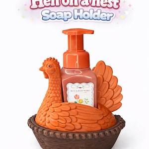 以下が含まれることがあります： オレンジ色のニワトリ型のソープホルダー、茶色の巣のベース付き。 ホルダーにはポンプボトルの石鹸が入っています。 テキスト「Hen on a nest Soap Holder」が上に表示されています。
