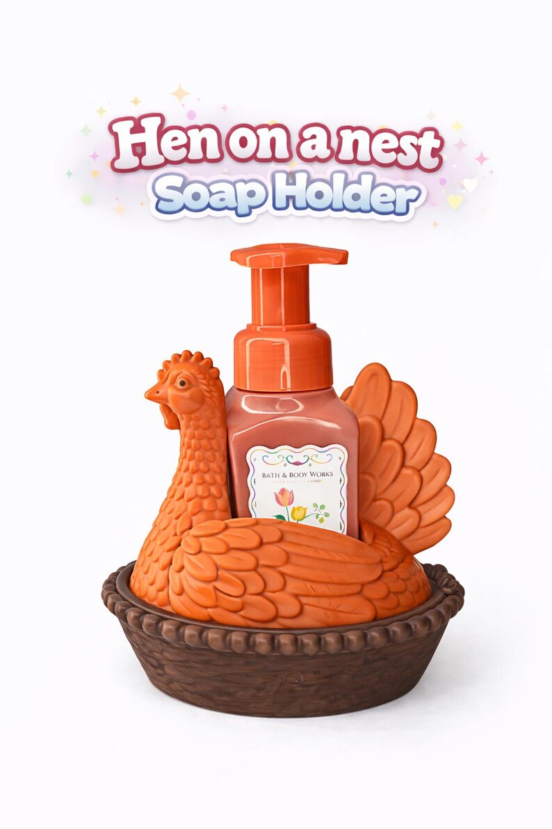 以下が含まれることがあります： オレンジ色のニワトリ型のソープホルダー、茶色の巣のベース付き。 ホルダーにはポンプボトルの石鹸が入っています。 テキスト「Hen on a nest Soap Holder」が上に表示されています。