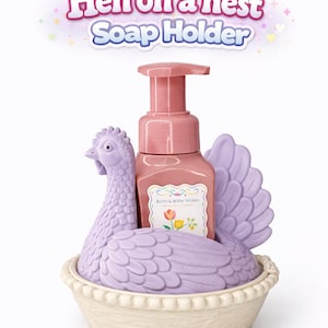 以下が含まれることがあります： ラベンダー色の鶏の形をした石鹸ホルダーとピンクのポンプボトルを備えた装飾的な石鹸ディスペンサー。ホルダーは巣のようにデザインされています。「Hen on a nest Soap Holder」のテキストが上に表示されています。