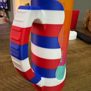 K&ouml;nnte beinhalten: Ein 3D-gedruckter Getr&auml;nkedosen-Griff mit einem rot-wei&szlig;-blauen Streifendesign. Der Griff ist so konzipiert, dass er um eine Dose in Standardgr&ouml;&szlig;e passt und einen bequemen Griff bietet. Der Griff ist aus Kunststoff.