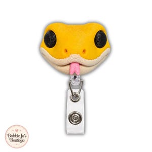 May include: A yellow snake head badge reel with large black eyes and a small pink tongue protruding from the mouth. The snake head is attached to a clear plastic strap with a metal snap button and a retractable cord mechanism for holding an ID card.