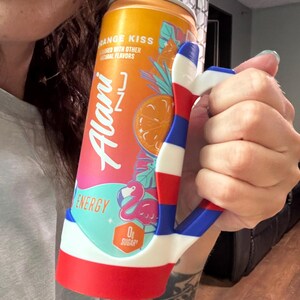 K&ouml;nnte beinhalten: Eine orangefarbene und rosa Alani Nu Energy-Drink-Dose, die in einem rot, wei&szlig; und blau gehaltenen handf&ouml;rmigen Halter gehalten wird. Die Dose tr&auml;gt den Text "Orange Kiss" und eine Flamingo-Grafik. Der Halter ist in einem patriotischen Farbschema gestaltet.