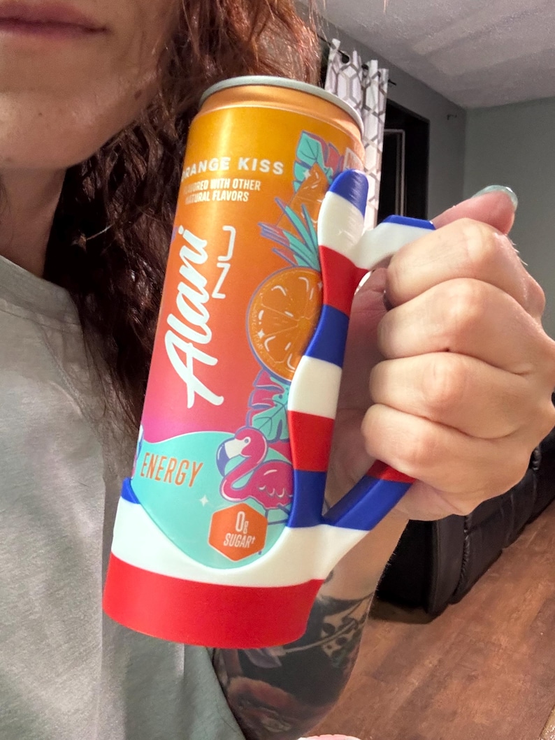 K&ouml;nnte beinhalten: Eine orangefarbene und rosa Alani Nu Energy-Drink-Dose, die in einem rot, wei&szlig; und blau gehaltenen handf&ouml;rmigen Halter gehalten wird. Die Dose tr&auml;gt den Text "Orange Kiss" und eine Flamingo-Grafik. Der Halter ist in einem patriotischen Farbschema gestaltet.