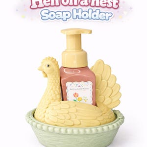 以下が含まれることがあります： 黄色いニワトリの形をした装飾的な石鹸ホルダー。薄緑色のバスケットの中にあります。 花柄のラベルが付いたピンク色の石鹸ディスペンサーが入っています。 テキスト「Hen on a nest Soap Holder」が上に表示されています。