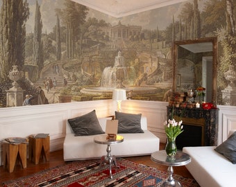 May include: A room featuring a large mural depicting a classical garden scene with a fountain, stairs, and figures. The room includes white sofas, silver side tables, a patterned rug, and wooden stools. A fireplace and mirror are also visible.