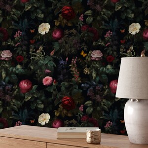Pu&ograve; includere: Una carta da parati floreale scura con un motivo ripetitivo di fiori rossi, rosa e bianchi, foglie verdi e farfalle. Una lampada in ceramica bianca con un paralume beige &egrave; posizionata su un tavolo di legno davanti alla carta da parati.