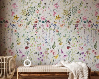 Könnte beinhalten: Eine hellbeige Wand mit einer floralen Tapete mit einem Aquarellmuster aus rosa, lila, gelben und grünen Blumen. Eine Holzbank mit einer weißen Keramikskulptur steht vor der Wand. Ein geflochtener Korb und eine Wolldecke liegen auf dem Boden.