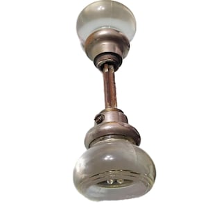 Op de afbeelding: Een vintage deurknopset bestaande uit twee heldere, bolvormige glazen knoppen verbonden door een centrale metalen spindel. De glazen knoppen zijn gemonteerd op doffe, zilverkleurige metalen voetstukken, met een zichtbare schroef op een kraag.