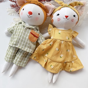 May include: Two handmade fabric dolls. One doll wears green and white plaid pajamas with a brown pocket. The other doll is dressed in a yellow floral dress. Both dolls have white faces, stitched features, and animal ears.