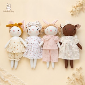 Pode incluir: Quatro bonecas de pano feitas à mão dispostas em fila. A primeira usa uma faixa de cabelo mostarda e um macacão floral. A segunda tem um vestido floral e faixa a combinar. A terceira usa um pijama rosa com gola de renda. A quarta é castanha com um vestido de renda branco.
