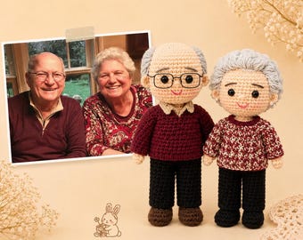 May include: Two handmade crochet dolls representing an elderly couple, standing next to a photograph of the same couple. The male doll wears a maroon sweater and black pants, while the female doll wears a patterned maroon sweater and black pants.
