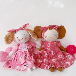 May include: Two handmade fabric dolls. One doll has a white face with a pink bow and a pink dress with a strawberry and dessert print. The other doll has a brown face with a pink bow and a pink dress with a daisy print. A pink yarn ball is next to the second doll.