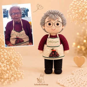 May include: A handmade crochet doll featuring gray yarn hair, black-rimmed glasses, a maroon long-sleeved shirt, and a cream-colored apron with green trim. The apron displays the embroidered text Las Cruces and a small red chili pepper graphic.