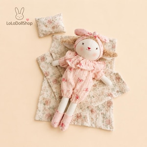 Puede incluir: Una muñeca de conejo de peluche con orejas largas, vestida con un pelele de flores rosas, un cuello con volantes a juego y una diadema. La muñeca viene acompañada de una pequeña almohada rectangular y una manta doblada con estampado floral.