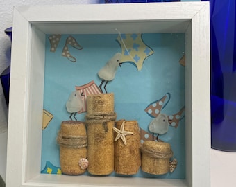 May include: A framed shadow box art piece featuring a beach scene. The artwork includes corks, twine, a starfish, and painted birds. The background is a light blue with illustrations of beach umbrellas and clothing. The frame is white.