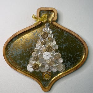 May include: A Christmas tree made from white, gold, and pearl buttons, set on a gold and green oval wooden tray. The tray has a wooden frame and a gold ribbon. The tree is decorated with buttons of various sizes and shapes.