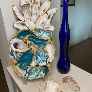 Puede incluir: Una escultura decorativa de piña hecha de conchas de ostra, pintada en turquesa y blanco con detalles dorados. También se exhiben un jarrón alto de cristal azul cobalto y varias conchas marinas.