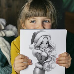 Puede incluir: Una ni&ntilde;a sostiene una ilustraci&oacute;n en escala de grises de una sirena con un gorro de Pap&aacute; Noel y un abrigo con ribete de piel. La sirena tiene el pelo largo y suelto y un copo de nieve en su abrigo. La ni&ntilde;a lleva una camisa amarilla de manga larga.