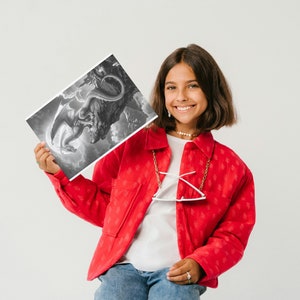 May include: A young person wearing a red jacket with a white shirt underneath and blue jeans is holding a black and white drawing of a dragon. The drawing is on a white piece of paper.