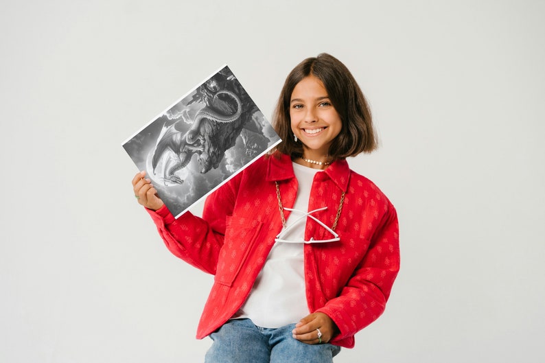 May include: A young person wearing a red jacket with a white shirt underneath and blue jeans is holding a black and white drawing of a dragon. The drawing is on a white piece of paper.
