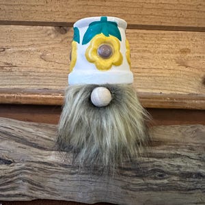 May include: A decorative gnome figure featuring a white ceramic pot base painted with yellow flowers and green leaves. The gnome has a long, shaggy grey faux fur beard and a smooth, round, light-colored wooden bead nose centered below the pot rim.