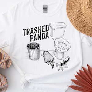 May include: White t-shirt featuring a black illustration of a raccoon, toilet, bin, and spilled bottle. The text "Trashed Panda" is above the graphic. The shirt is a Gildan brand.