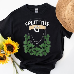 Pode incluir: Camiseta preta com o texto "SPLIT THE G" em branco e dourado. O gráfico apresenta um copo de cerveja stout estilizado com uma barba de trevo. Um chapéu de palha e girassóis estão no fundo.
