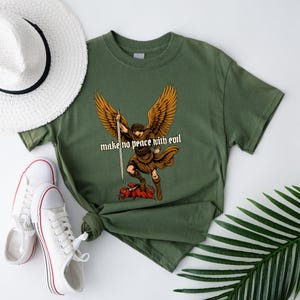 May include: Olive green t-shirt featuring a graphic of an angel with large wings, holding a sword, and standing on a red creature. The text "make no peace with evil" is printed below the angel. White sneakers and a sun hat are also in the image.