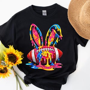 May include: Black t-shirt featuring a colorful graphic of a football with bunny ears. The football is red and white with colorful dripping paint. The bunny ears are pink, blue, and yellow. A sunflower and straw hat are in the background.