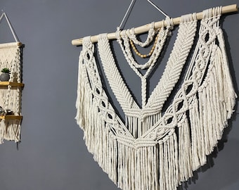May include: A white macrame wall hanging with wooden dowel and beaded accents. The hanging features a geometric design with fringe details.