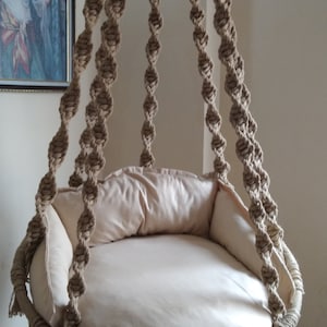 May include: A hanging macrame chair with a beige cushion. The chair is made of natural rope and has a circular shape. It is suspended from the ceiling by a metal hook.