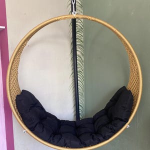 May include: A gold-colored round hanging chair with a black cushion. The chair is suspended from a black metal stand.