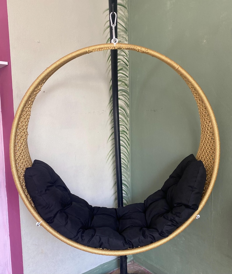 May include: A gold-colored round hanging chair with a black cushion. The chair is suspended from a black metal stand.
