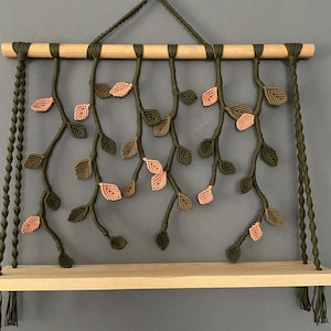 May include: A macrame wall hanging with a wooden shelf. The macrame is made with green and pink yarn and features a leaf design.