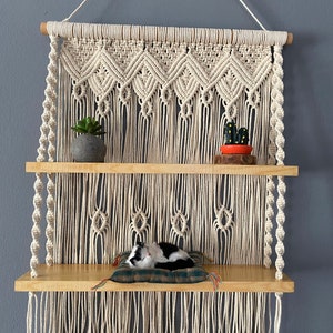 Puede incluir: Una estantería de madera de dos niveles con una macramé colgando encima. La estantería está decorada con una pequeña planta en maceta, un cactus y un gato blanco y negro durmiendo en una almohada.