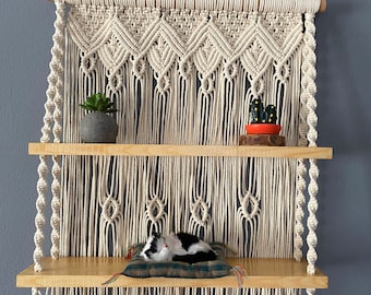 May include: A two-tiered wooden shelf with a macrame hanging above. The shelf is decorated with a small potted plant, a cactus, and a black and white cat sleeping on a cushion.