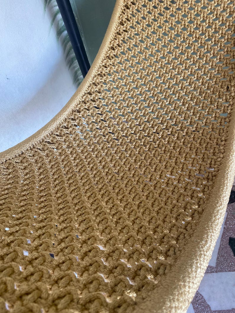 May include: A close-up of a woven hammock chair made of a light brown rope. The chair is made of a tight weave and has a smooth, rounded edge.