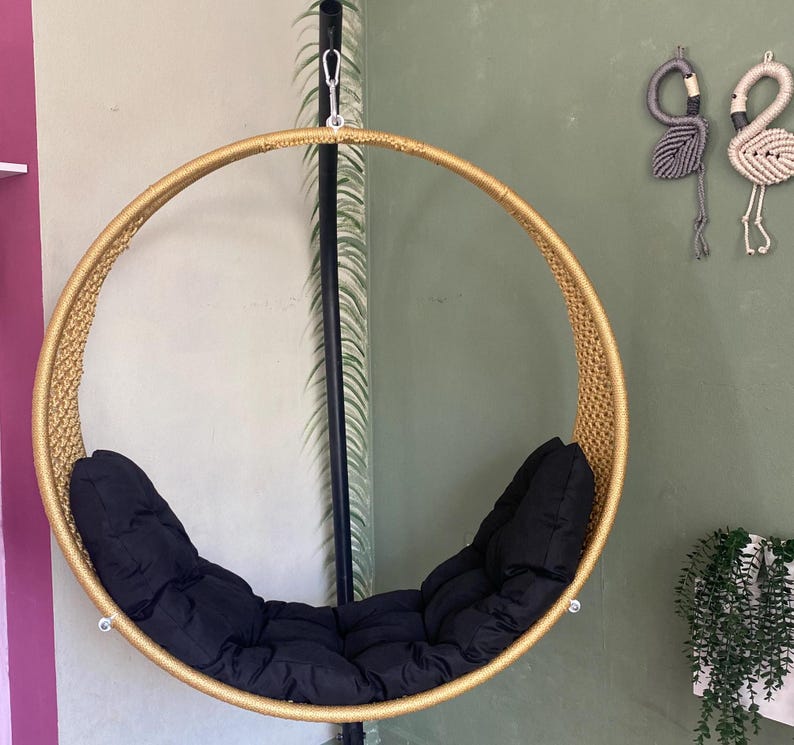 May include: A gold-colored round hanging chair with black cushions. The chair is suspended from a black metal stand.